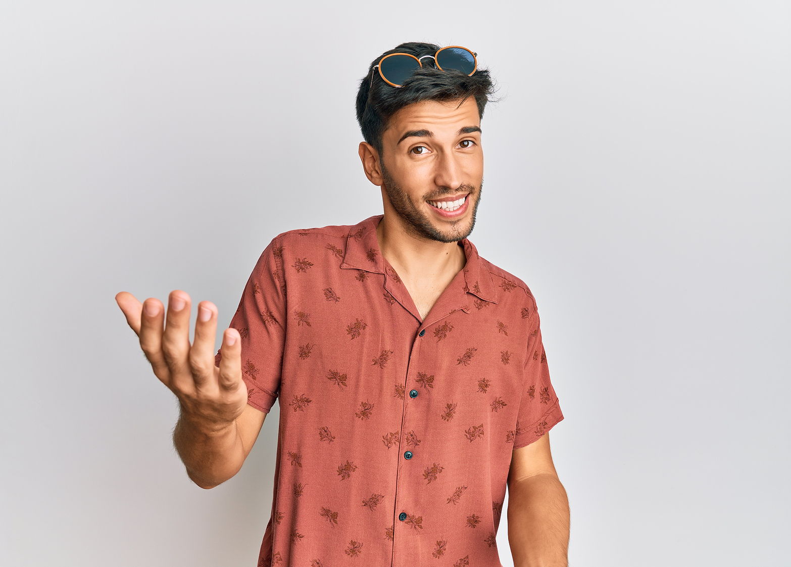 Photo of man wearing a red short sleeve and glasses on his head smiling reaching his hand out to you with white background.