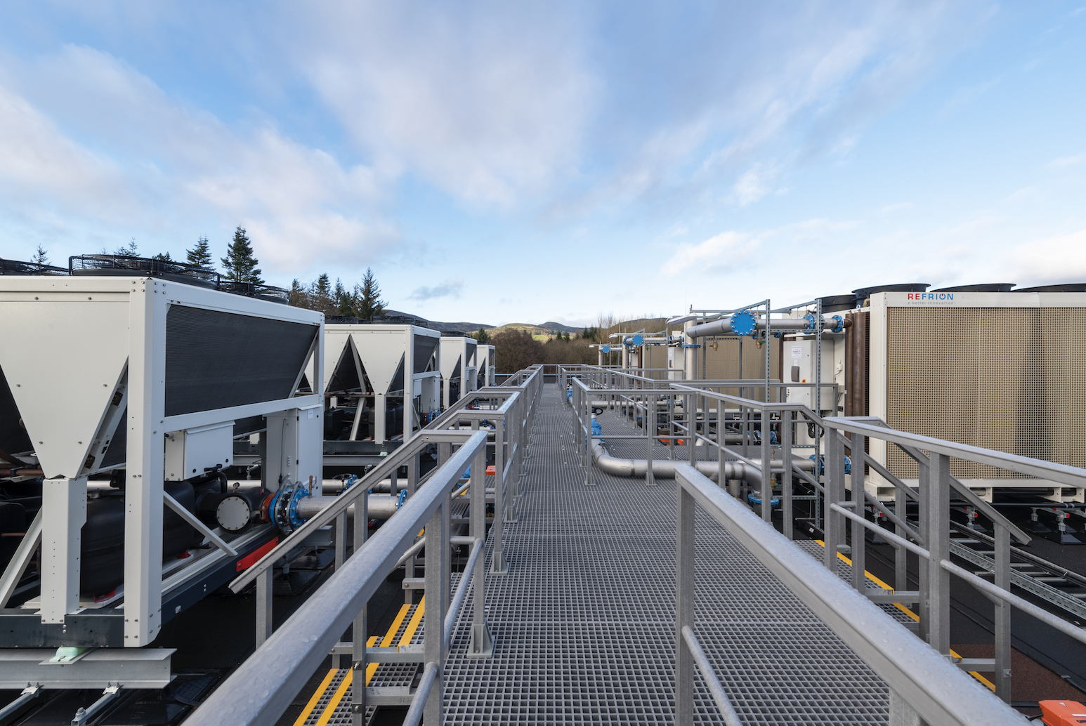 View of industrial HVAC units and cooling systems on a rooftop, with metal walkways, pipes, and railings under a partly cloudy sky and surrounded by trees in the background.