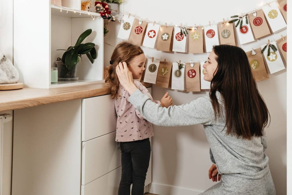 A mother gently tucks her daughter’s hair behind her ear while they smile at each other in a cozy kitchen decorated with a handmade Advent calendar made of paper bags and festive ornaments.