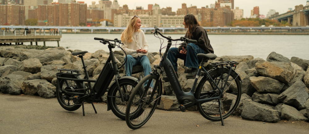 two women sitting on rocks over the hudson river behind their electric city bikes in new york city