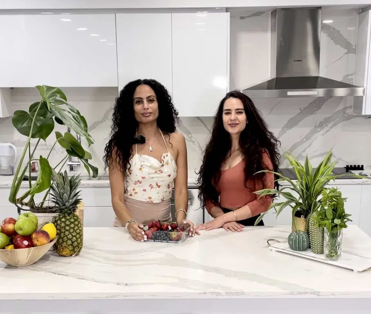 Two women stand behind a kitchen island with berries and plants, with a modern range hood and white cabinetry behind them.