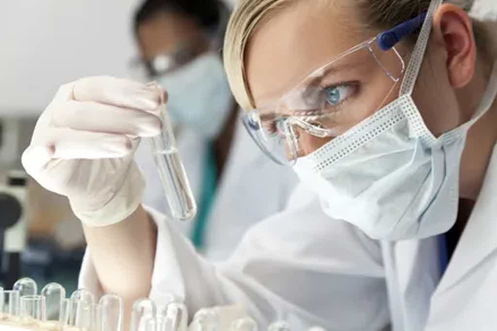 Scientist wearing protective gear closely examining a test tube in a laboratory setting with another researcher in the background.