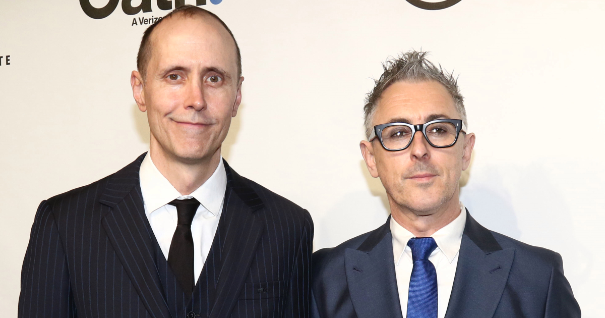 Image of Alan Cumming with his husband Grant Shaffer being photographed at a movie screening. Both are wearing dark colored suits and are smiling.