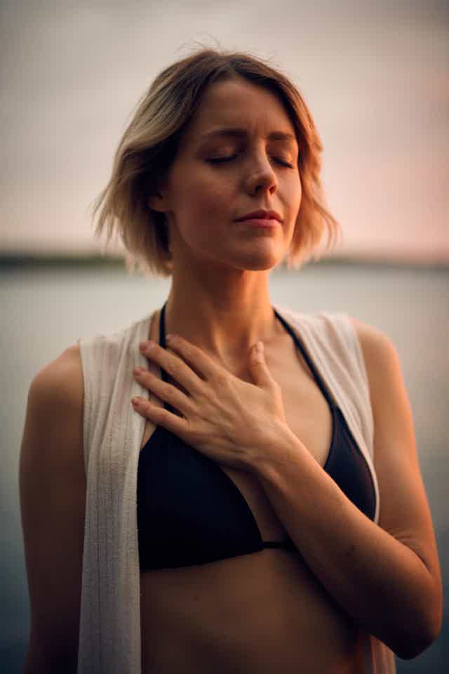 Woman with closed eyes and hand on chest practices mindfulness outdoors at sunset, expressing calm and self-awareness.