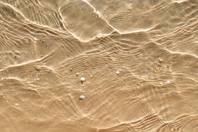 At the beach the shallow clear water shows the tiny pebbles on the sand.