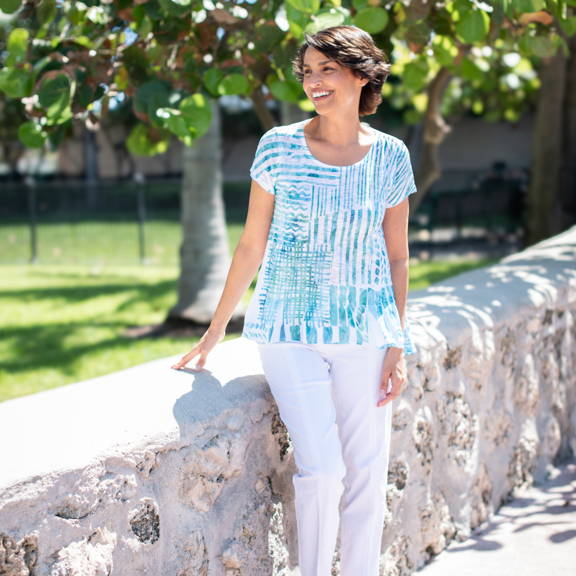 A woman wears a striped blue top and white pants as she stands in a sunny park.