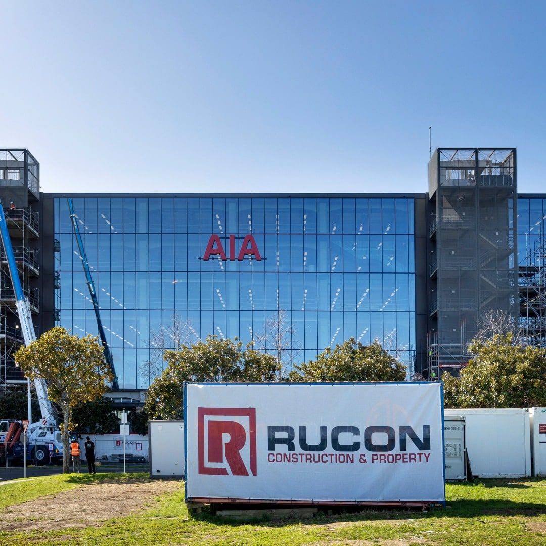 A modern glass office building with AIA in red letters on the facade. In front, theres a large RUCON Construction & Property sign and construction equipment visible on both sides. Trees and grass are in the foreground.