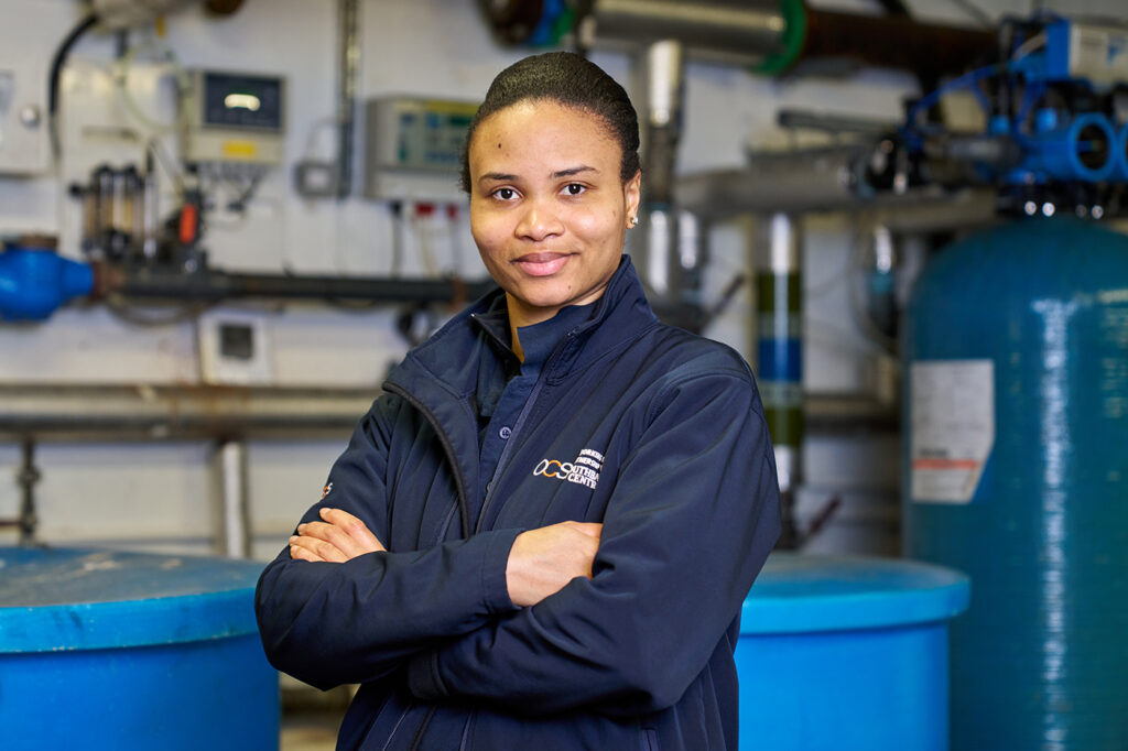 A woman wearing a dark blue jacket stands confidently with her arms crossed in an industrial setting, surrounded by large blue tanks and various pipes and control panels.