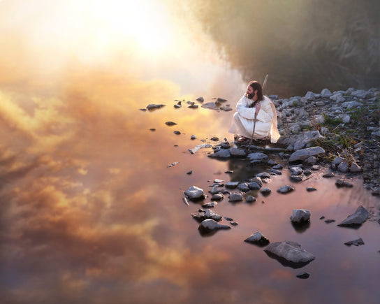 Jesus courched on a rocky shore looking out a lake that reflects the purple sky.