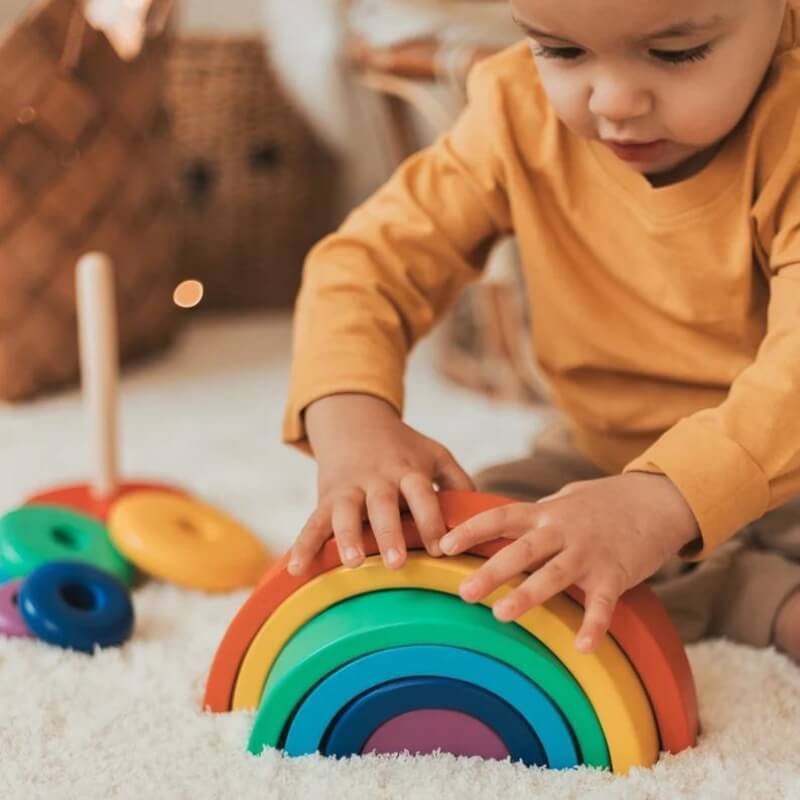 Un tout-petit en chemise jaune joue avec un jouet coloré et écologique en bois InvenToy Montessori Rainbow, en empilant ses pièces, sur un tapis blanc doux à l'intérieur.