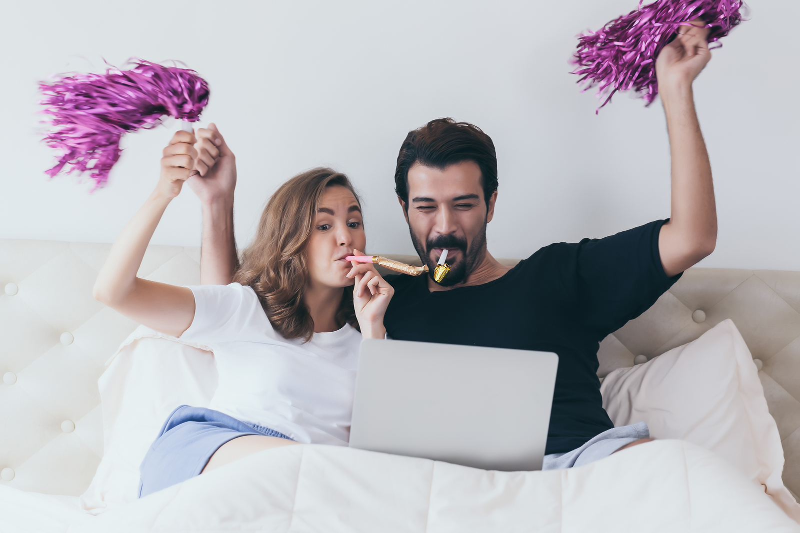 An attractive man and woman lying on bed with a laptop in front of them with fun party things in their hands.