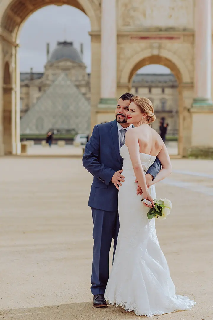 Photo de la mariée et du marié devant le musée du Louvre, se souriant mutuellement tandis qu'il la serre dans ses bras par derrière.