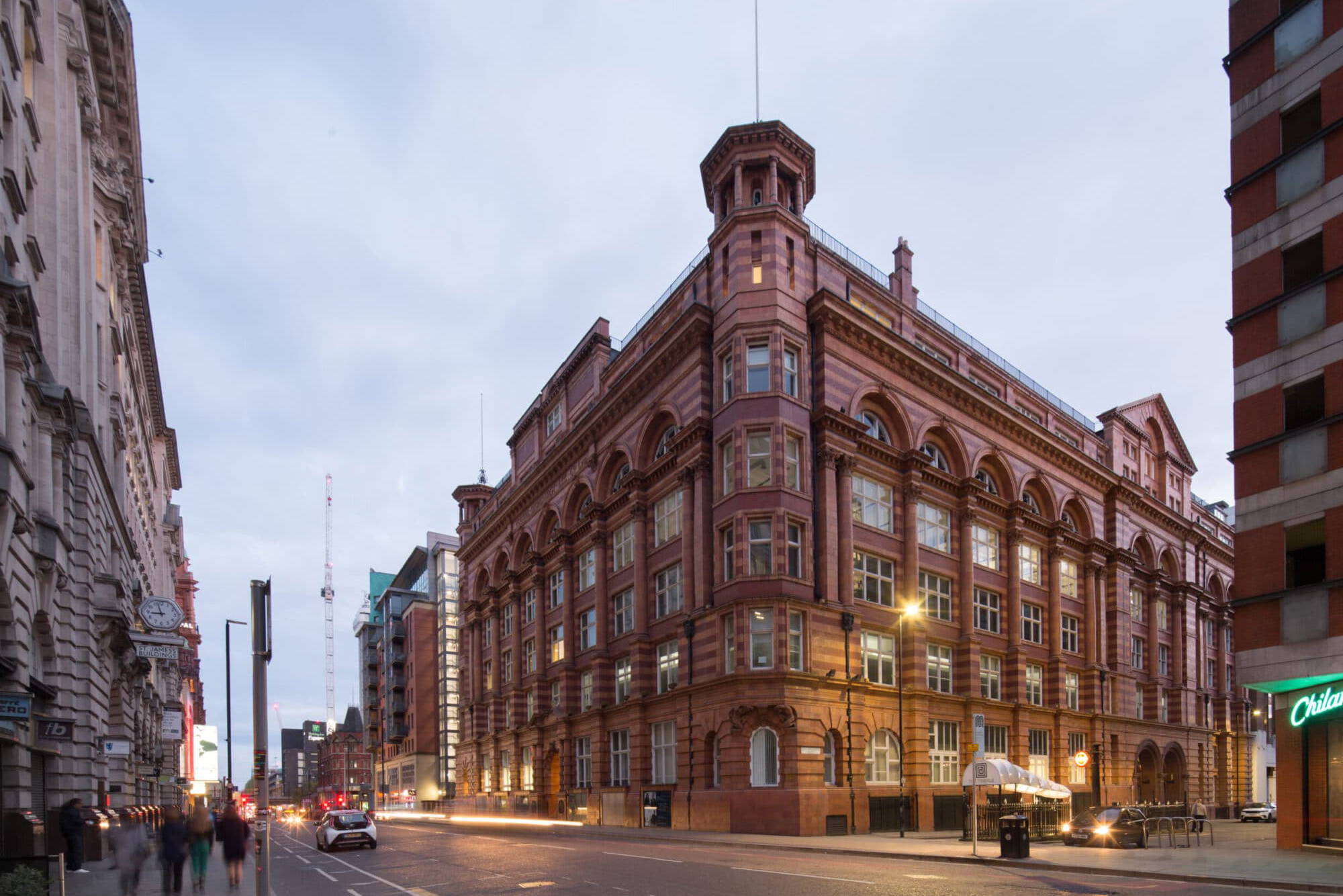 An image of the exterior of the historic Tootal buildings in Manchester from a 3/4 perspective. There is a long exposure on the image to blue cars and pedestrians walking past and it is just moving to the evening.