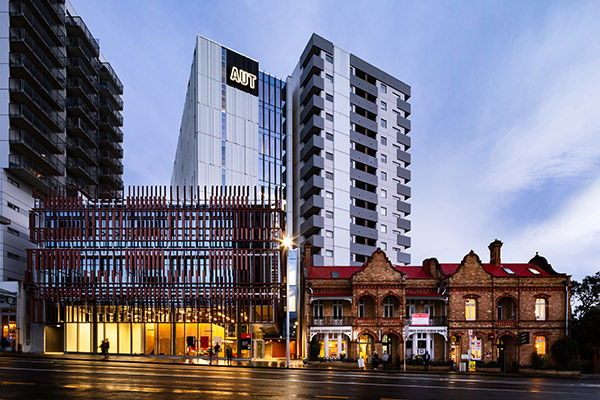 A modern glass building with AUT signage stands beside tall apartment blocks and a historic brick building with red roofs, all facing a wet street at dusk.