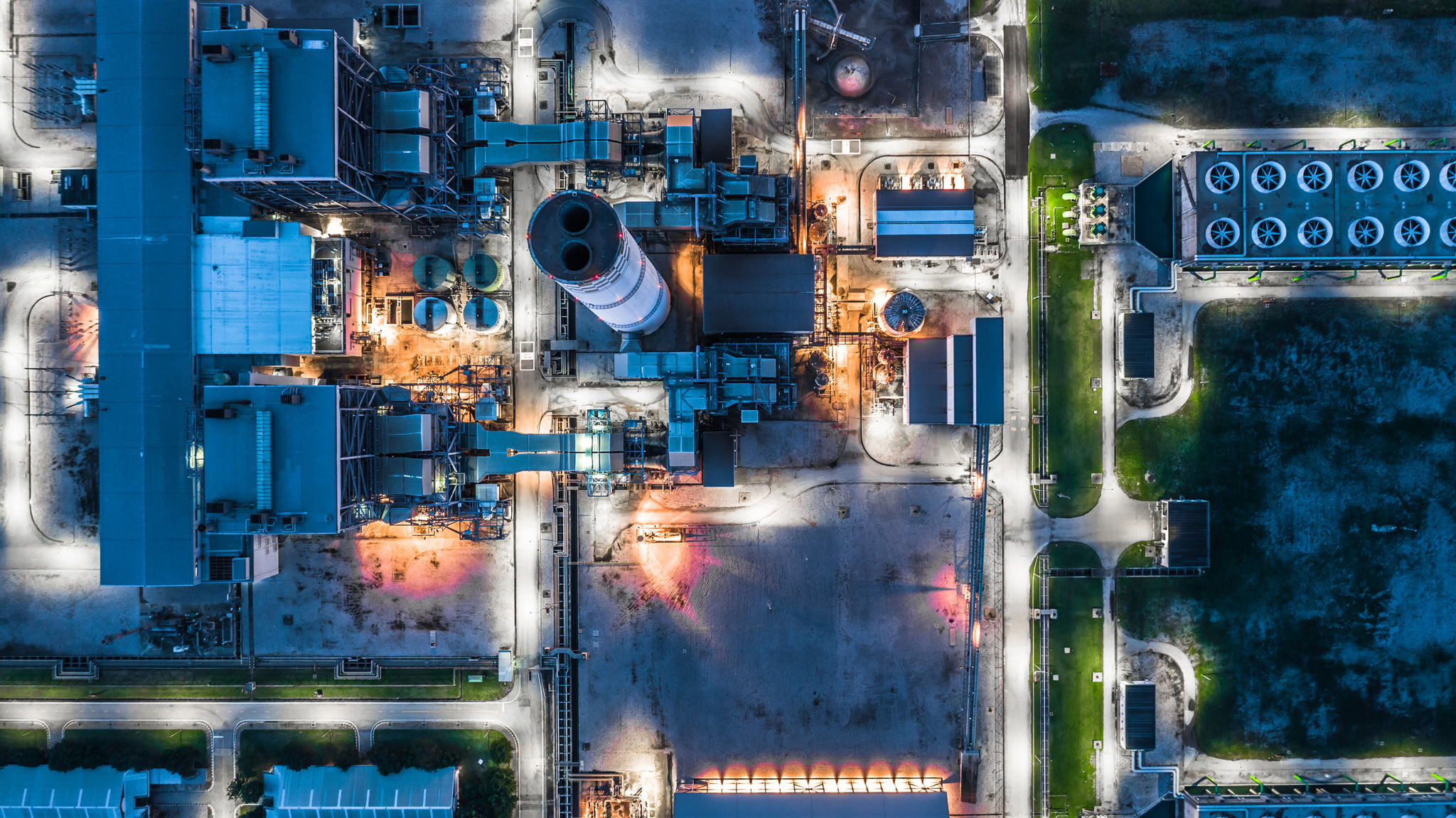 Aerial view of an industrial facility at dusk, with illuminated buildings, machinery, a tall chimney, and surrounding roads, creating a grid-like pattern. Some green areas are visible along the perimeter.
