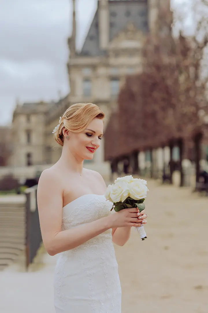 Photo de la mariée dans sa robe de mariée blanche. Elle tient des fleurs, les regarde et sourit.