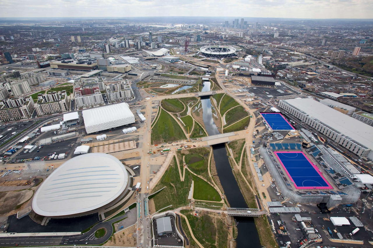 Aerial view of a large urban park with sports venues, stadiums, blue hockey fields, rivers, bridges, and surrounding city buildings under a cloudy sky.