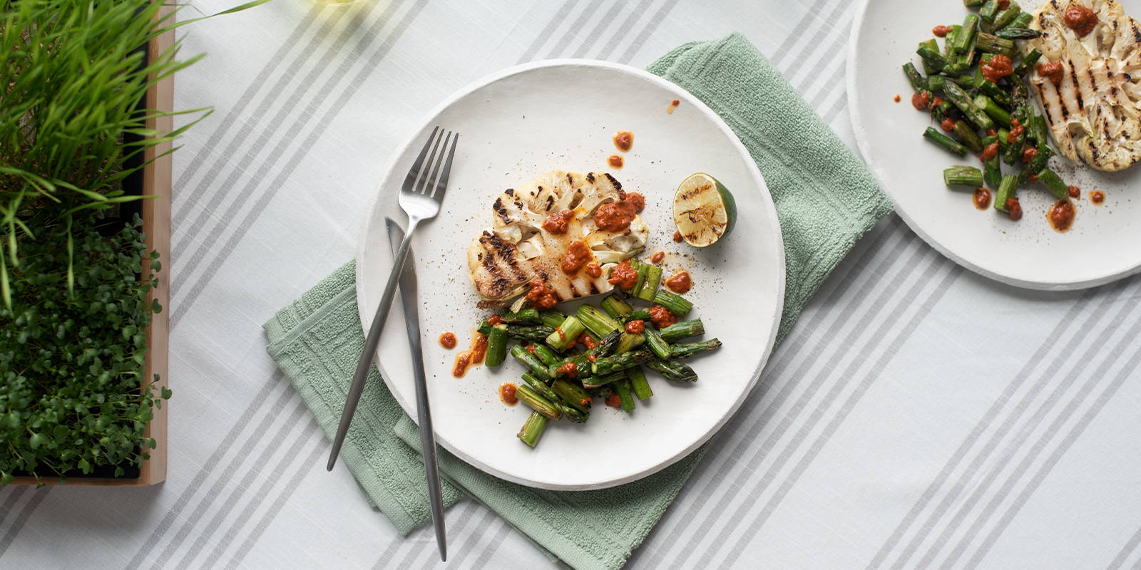 Two plates of grilled cauliflower and grilled asparagus, garnished with harissa and lime