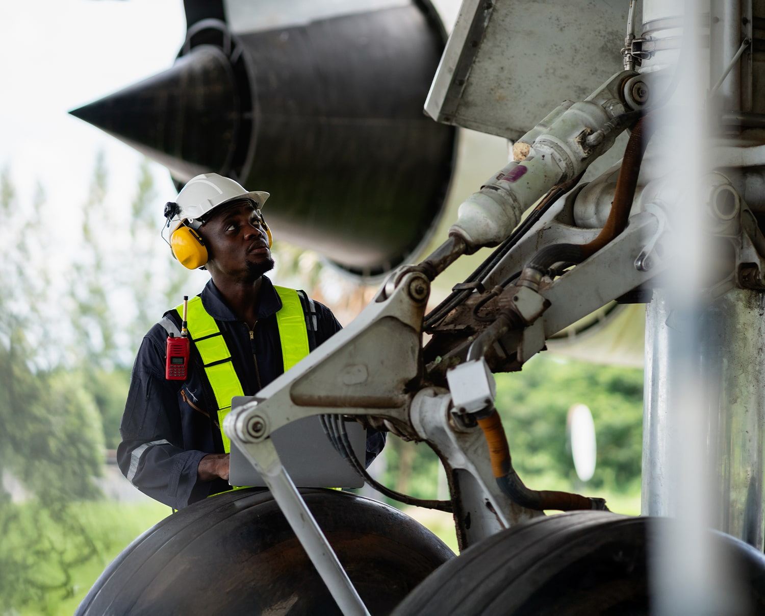 A male engineer wearing a safety vest, hard hat, and ear protection inspects the landing gear of an aircraft outdoors. Trees and greenery are visible in the background.