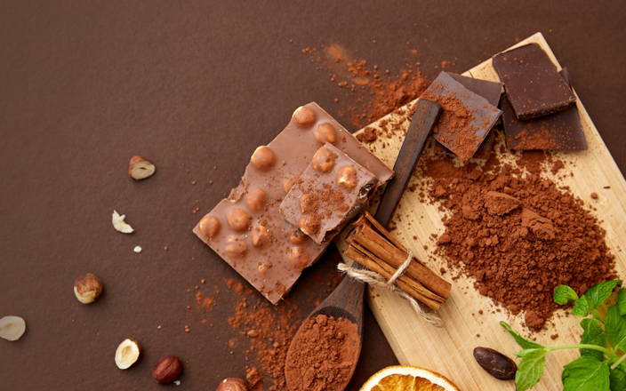 A cutting board with cocoa powder, cinnamon sticks, crushed hazelnuts, and a chocolate bar on it and scattered around for Confetti's Virtual Chocolate Tasting Team Building