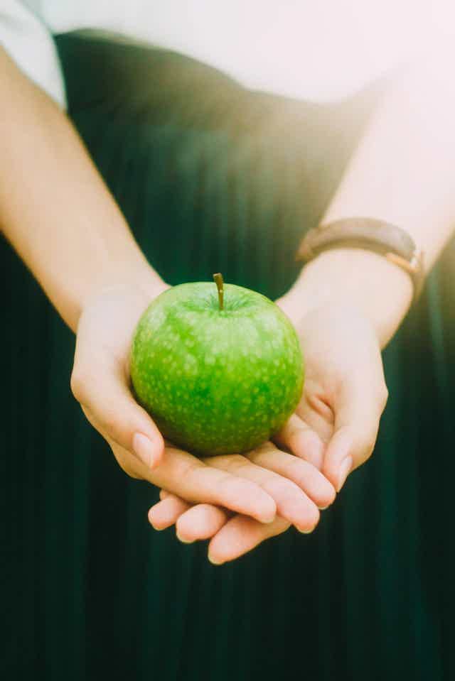 Person holding a fresh green apple with both hands, standing outdoors in soft natural light.
