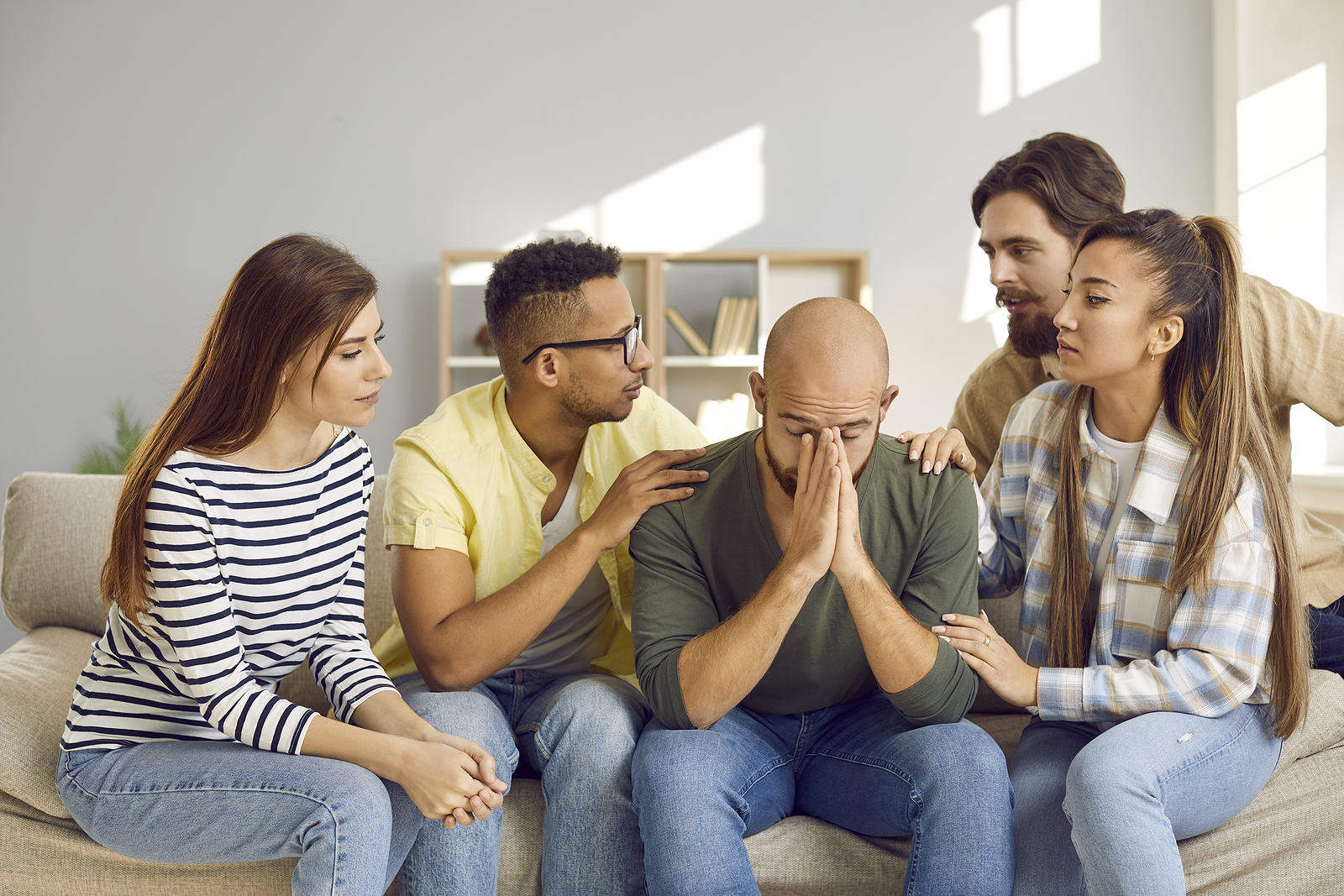 A group of friends sit next to their friend comforting him while he sits frustrated with his hands on his face.