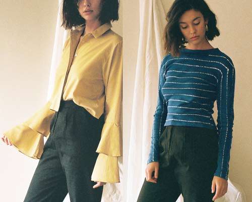 Woman wearing yellow frill sleeve blouse tucked into black high waisted trousers and woman wearing organic cotton wide and high neck long sleeve tee from sustainable fashion brand Leo Strange