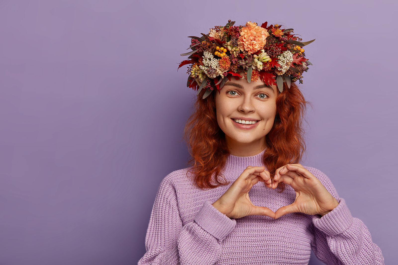 Image of a red haired woman with a large floral arrangement on top of her head. wearing a purple turtle neck and is making a heart with both her hands.