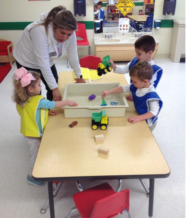 Three Primrose toddlers play with sailboats floating in a tray of water as their teacher talks to them