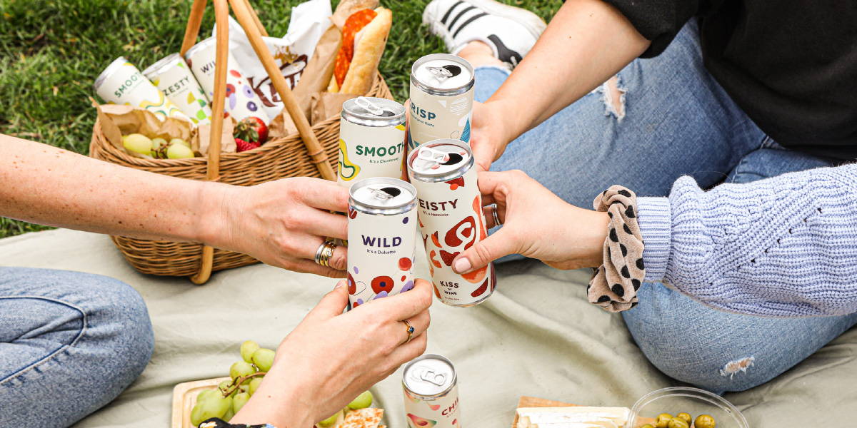 Picnic basket surrounded by light snacks with Kiss of Wine Sauvignon Blanc, Riesling and Rose cans. 