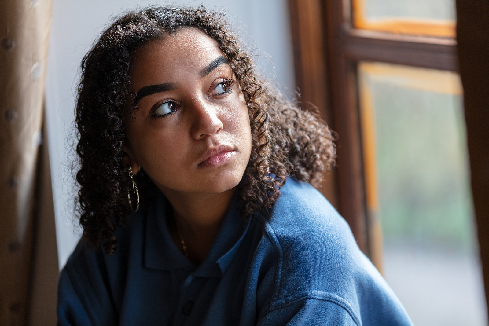 Beautiful mixed race African American girl teenager female young woman sad depressed or worried looking out of a window.