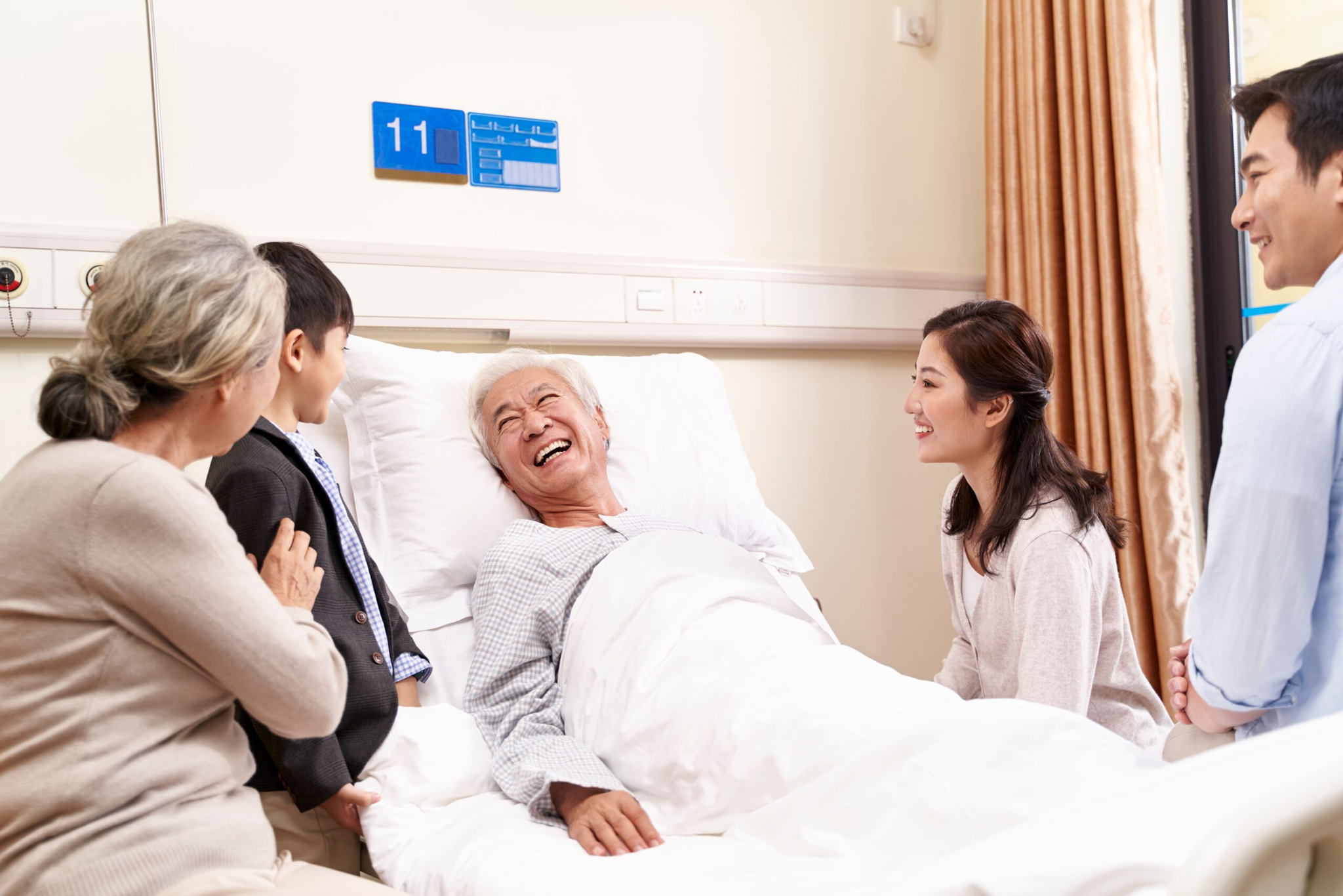 An elderly man lies in a hospital bed smiling and laughing, surrounded by four family members, including a child, in a bright hospital room.