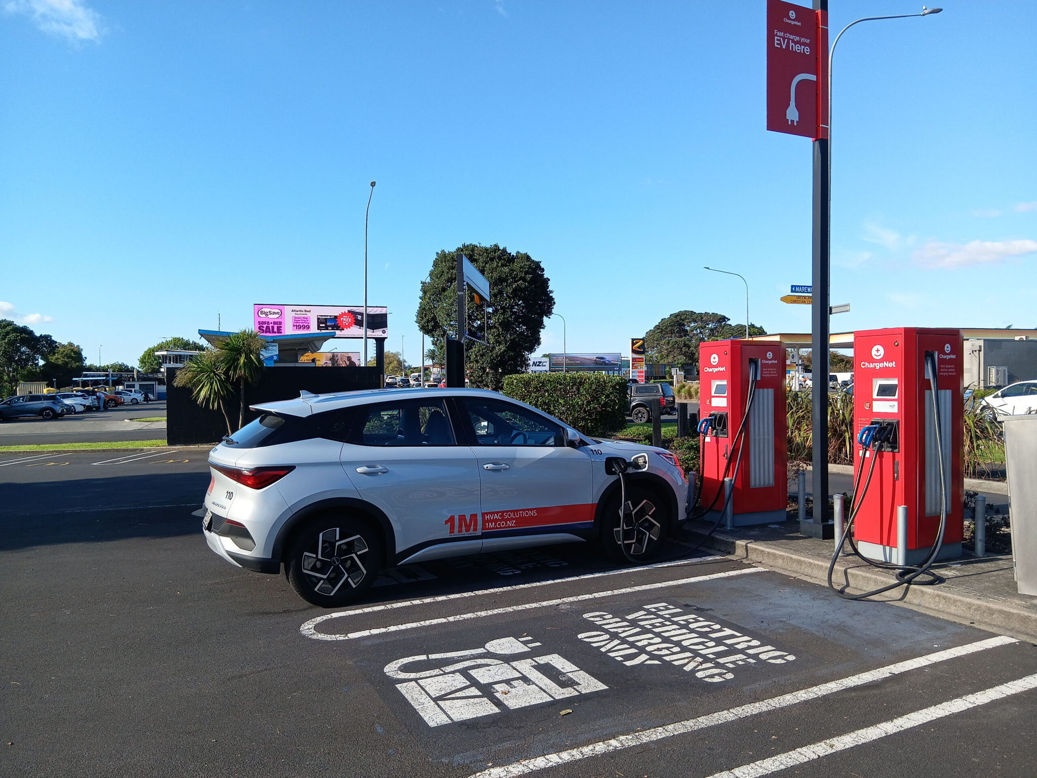 A white electric car is parked and charging at a public charging station in a parking lot. The station has red chargers and signs indicating it is for electric vehicles only. Trees and billboards are visible in the background.