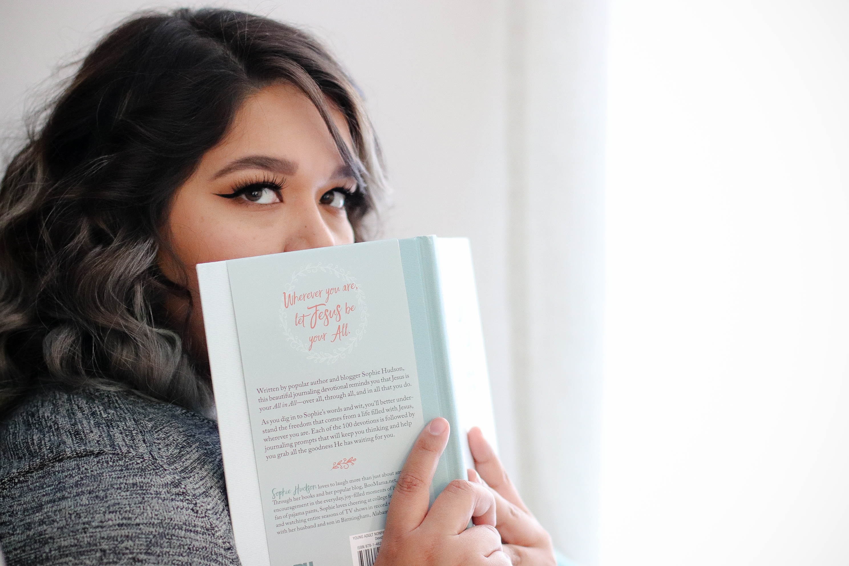 A woman hides her face behind a book while looking at the camera.