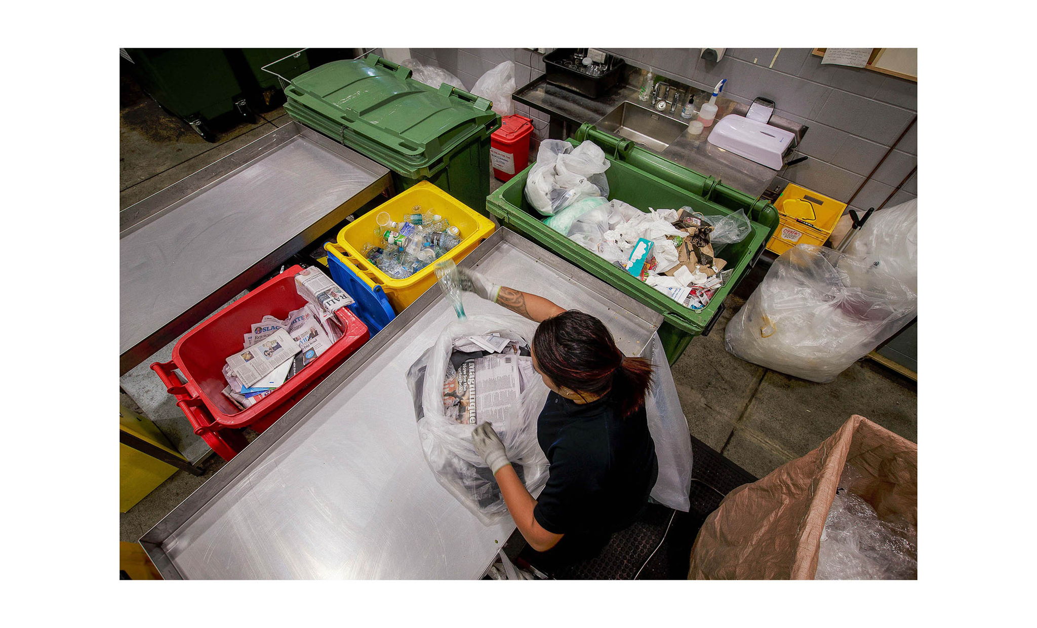 A person sorts recyclable materials such as paper, plastic bottles, and other waste into colored bins at a recycling facility, viewed from above. The area has stainless steel tables and various containers.