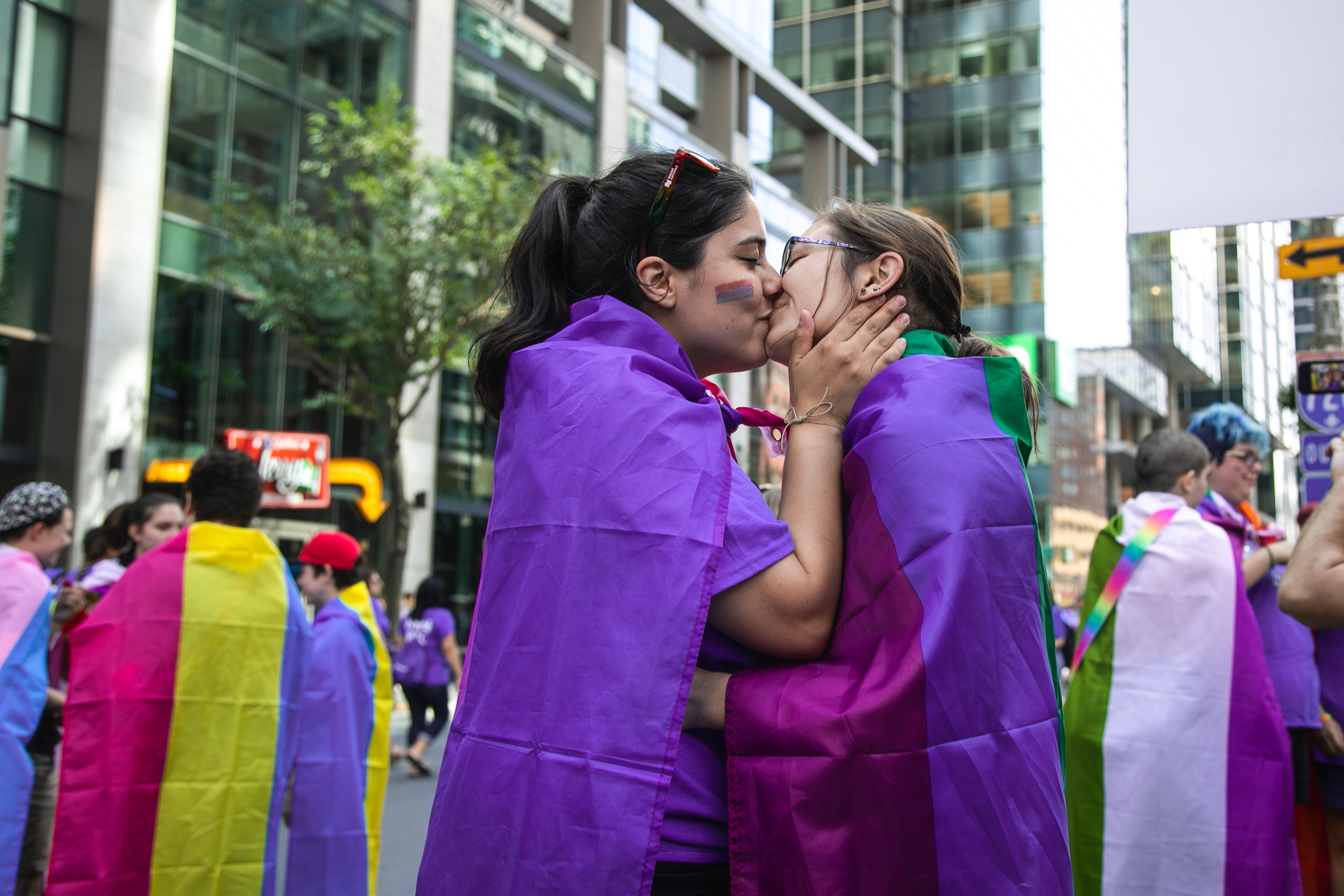 Two young women with long hair in the middle of the street during a pride parade, both wearing the bi flags as blankets kissing while one has her hands on the other girls' face.