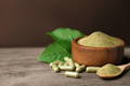 green powdered wheat grass in a wooden bowl, with several green wheat grass capsules scattered on the table