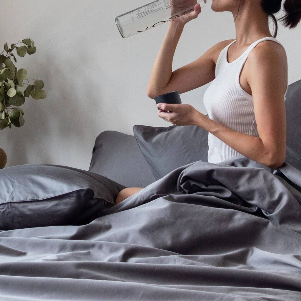 Girl drinking water while sitting on Weavve's grey cotton bed sheets