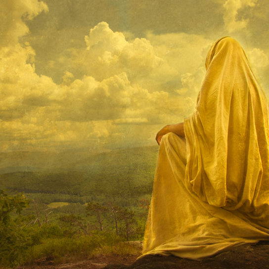 Jesus in a yellow robe sitting on a hill and looking out at a lush landscape.