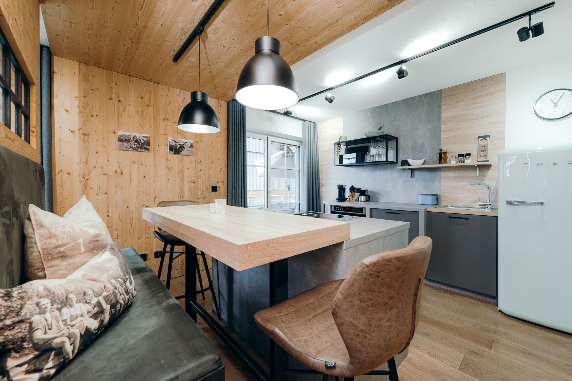 Im Vordergrund ein hölzerner Essbereich mit grauer Sitzbank, im Hintergrund eine moderne Küche mit hellem Kühlschrank.