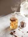 A clear glass tea pot pours steaming hot water into a carafe filled with dark colored herbs