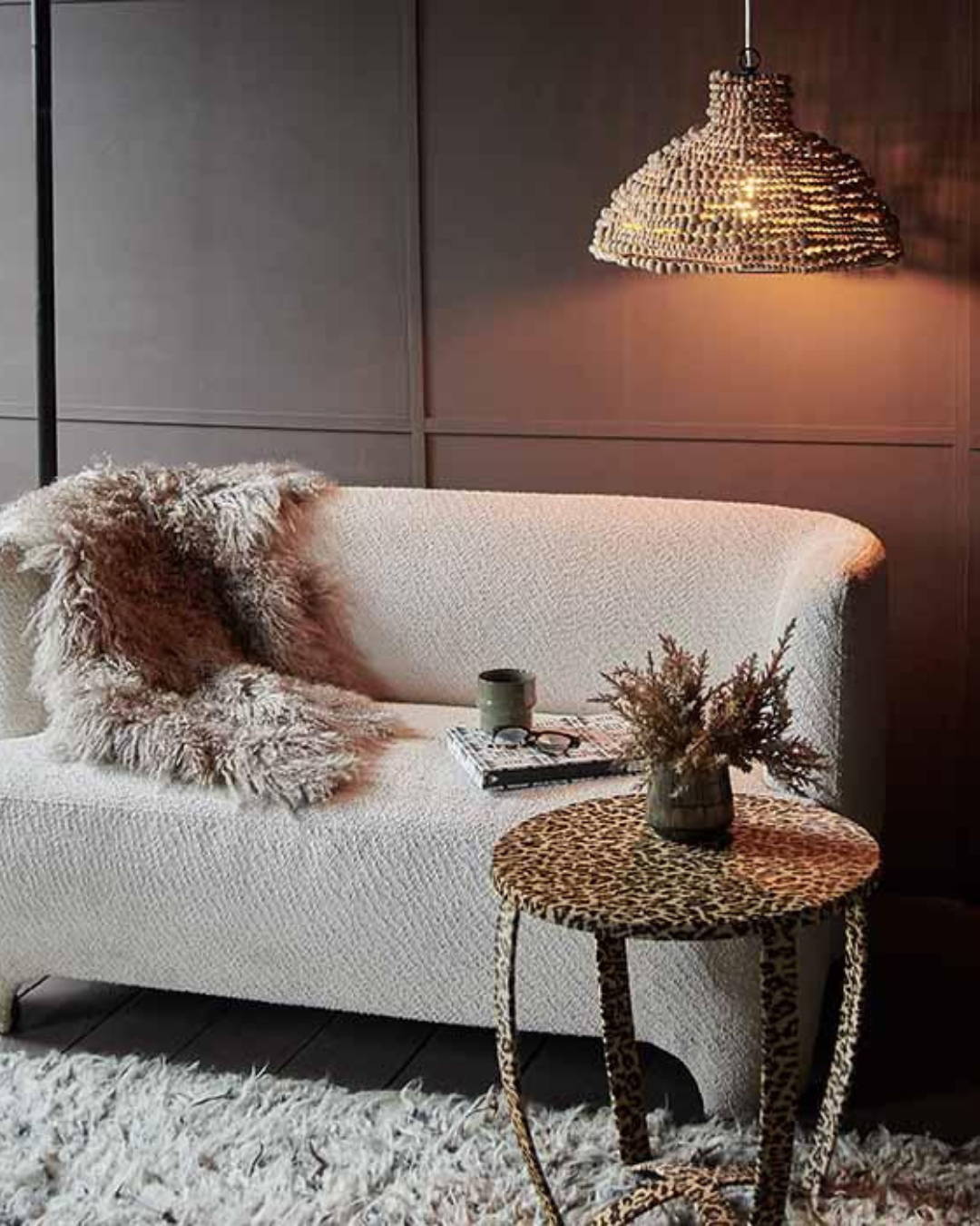 A curved cream sofa draped with a sheepskin throw next to a table that has a leopard print pattern and housing a plant with a beaded chandeliier
