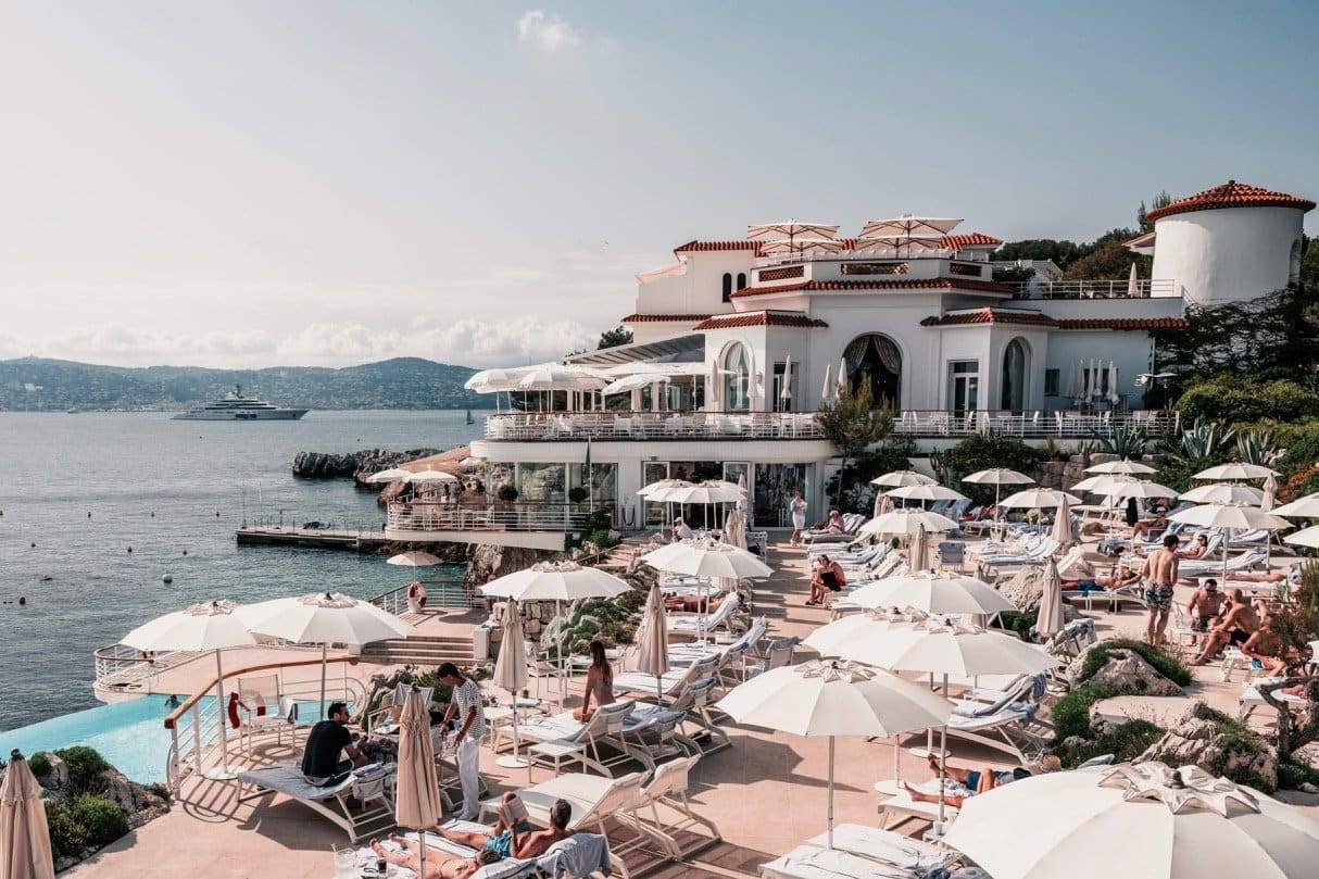 Eden Afternoons by Stuart Cantor - A photograph of Hotel du Cap, iconic oceanfront luxury hotel, captured with holidaymakers relaxing beneath white sun umbrellas in the foreground with glittering ocean in the distance.