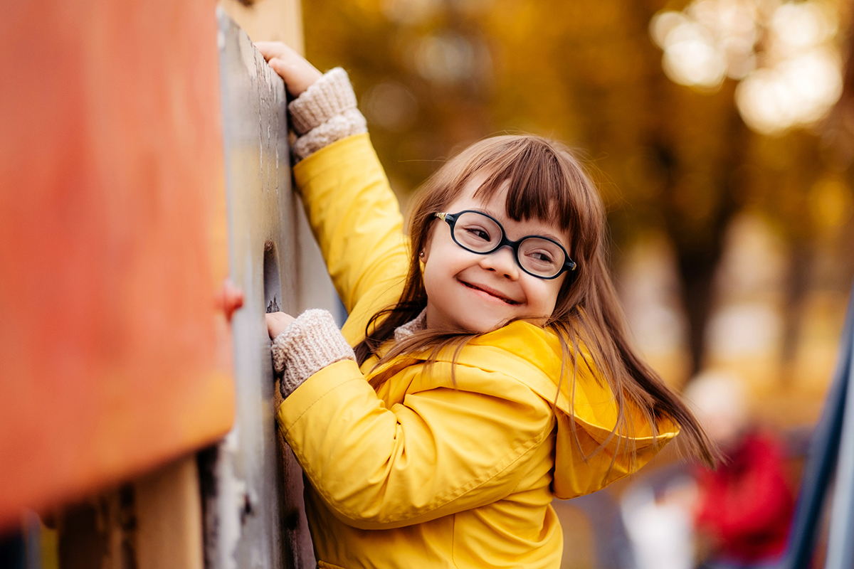 A young girl with glasses and long brown hair, wearing a yellow coat, smiles while climbing outdoor playground equipment. Autumn trees with yellow leaves blur in the background.
