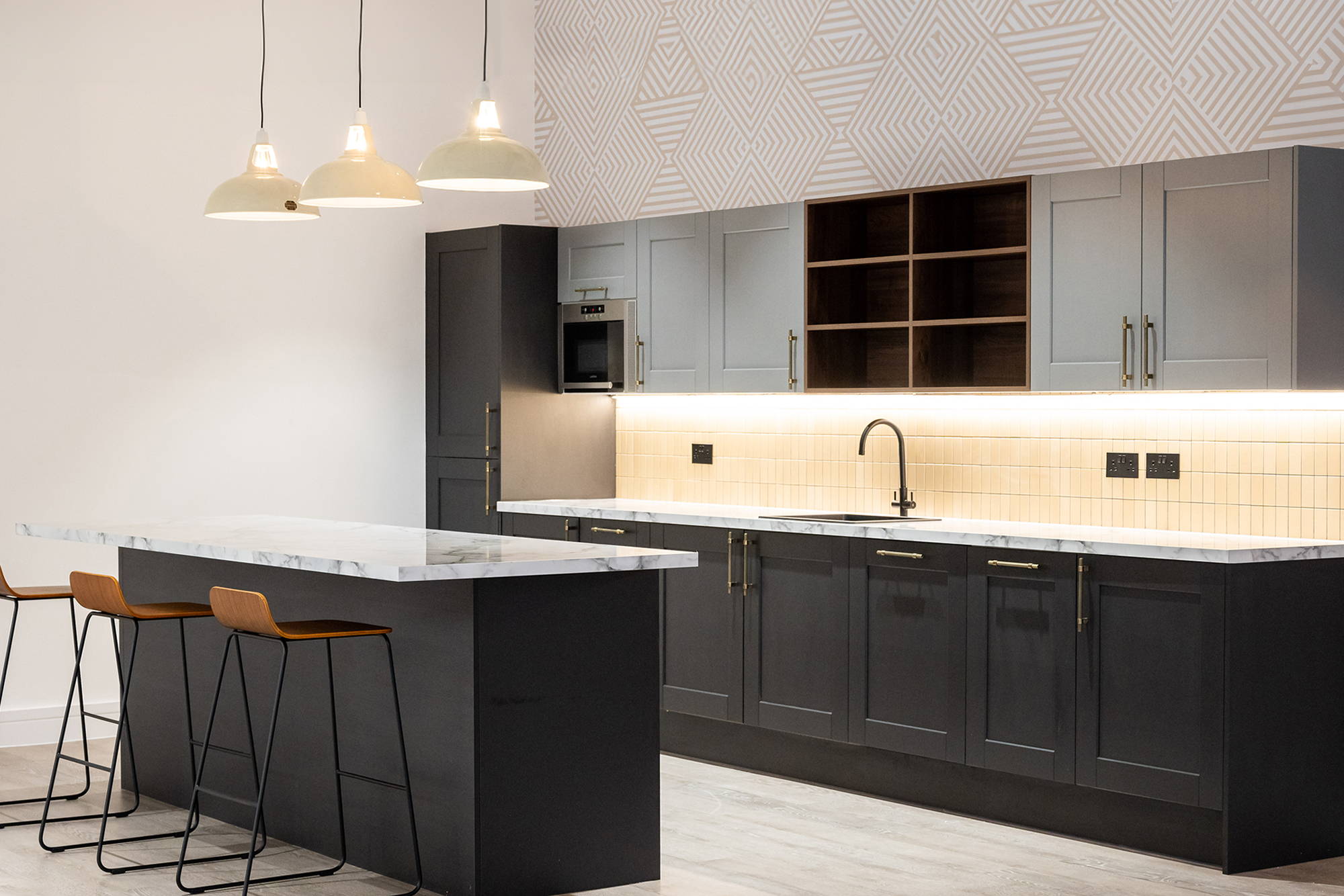 Three Large Yellow Pendant Lampshades hang in a row over a tiled bar, separating the kitchen area behind it, with Yellow barstools on the other side. On the back walls is hand-painted artwork in bright colours.