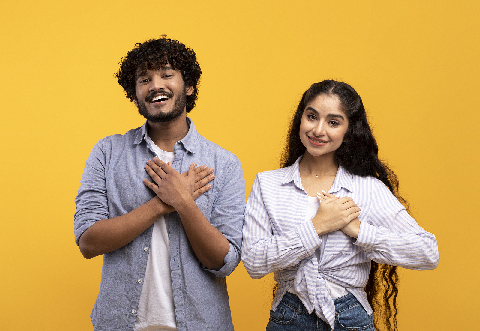 Thankful indian couple keeping both palms on chest, smiling at camera, expressing appreciation and kindness.