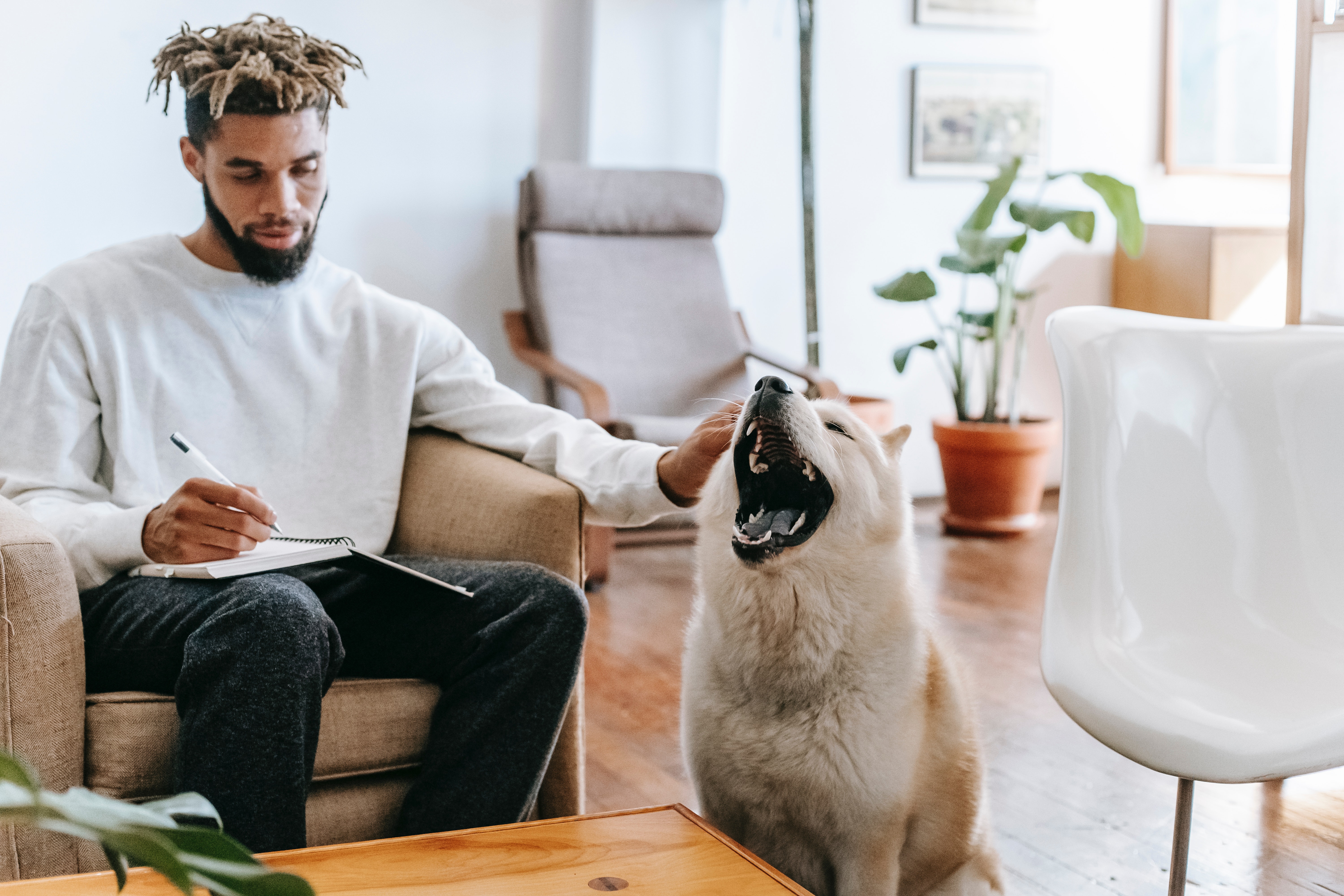 An attractive black man with dreads sits on his couch at journaling his thoughts while petting his yawning dog and smiling.