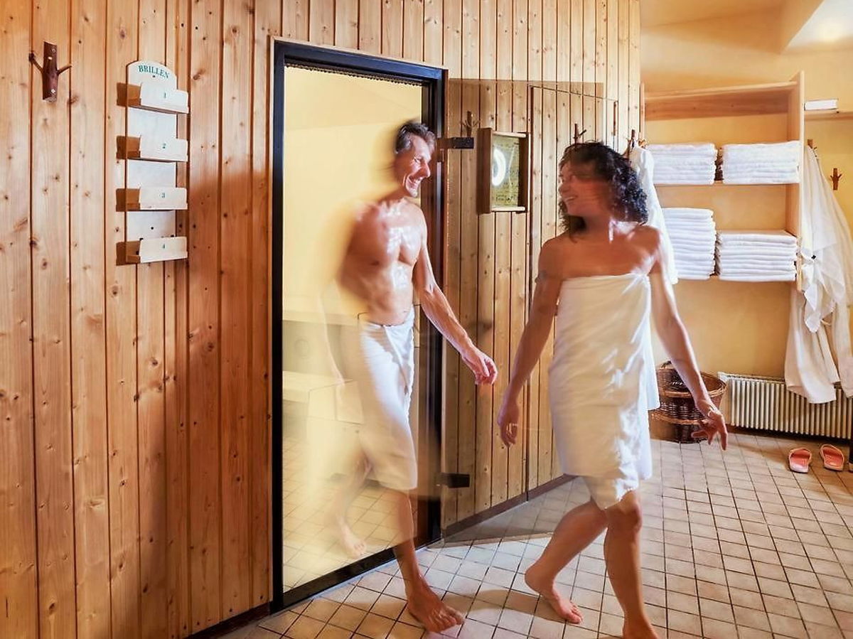 Ein Pärchen läuft aus einer Sauna. Beide mit weißen Handtüchern bedeckt. Wandfassade aus Holz