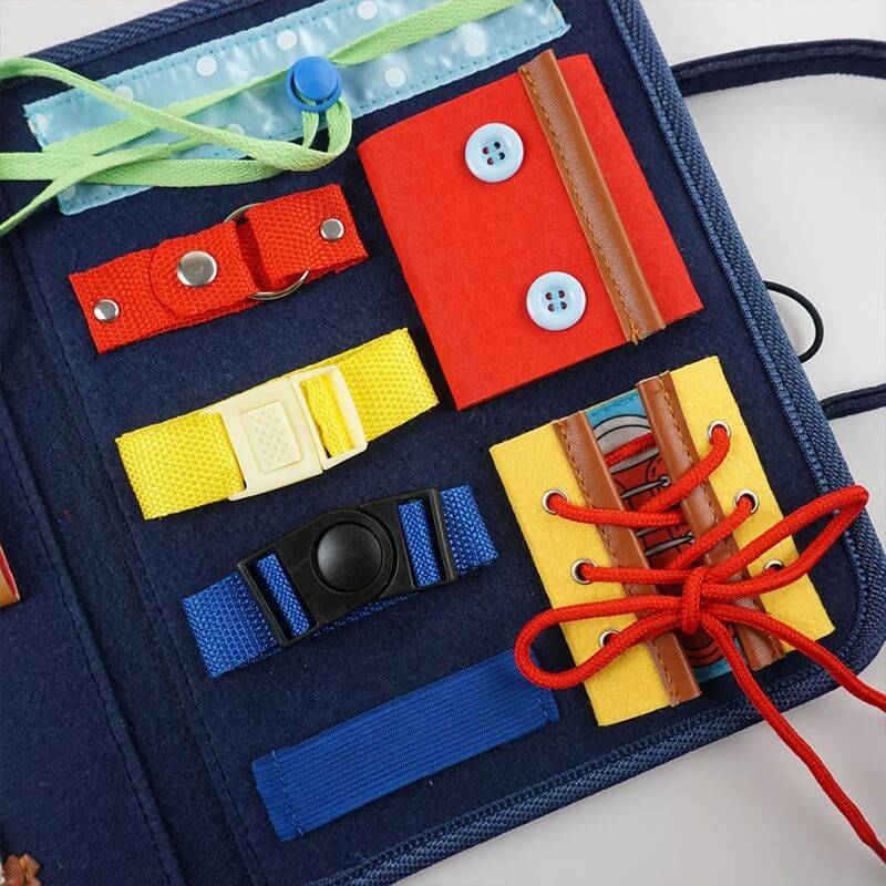 Un tableau d'activités Montessori InvenToy pour enfants présentant diverses textures et éléments de développement de compétences comme des boutons, des boutons-pression, une fermeture éclair et des lacets sur un fond en tissu bleu.