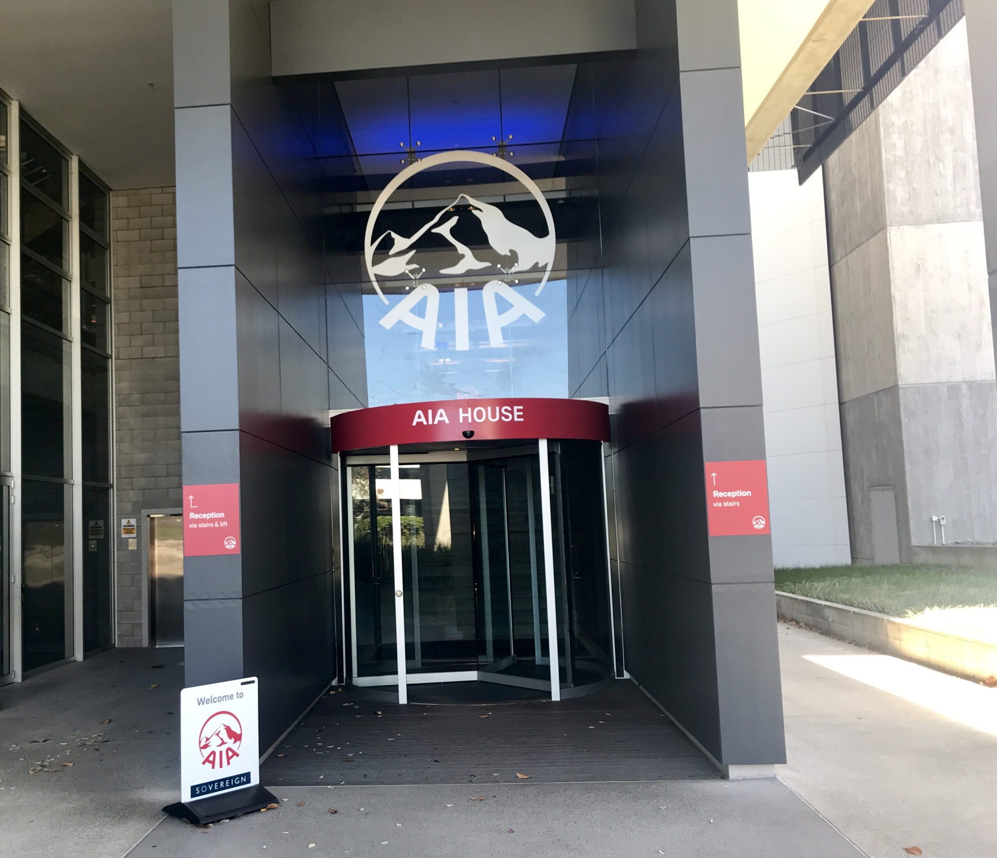 Entrance to a modern building labeled AIA House with a glass revolving door, AIA logo above, and welcome signs outside. The area is clean and well-lit with a mix of concrete and glass architecture.